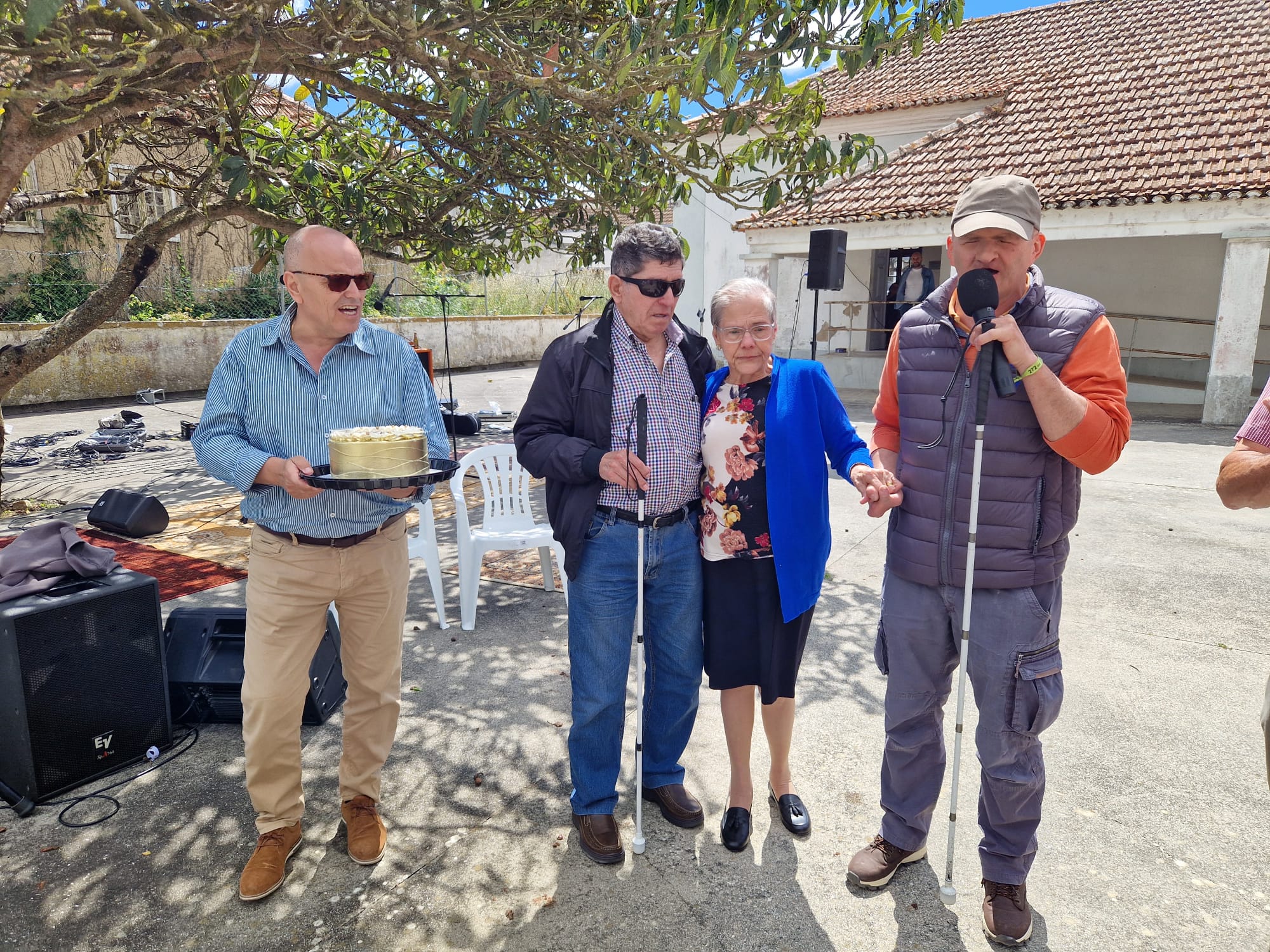 A imagem mostra quatro pessoas no aniversário da Associação Cabra Cega em ambiente ao ar livre. À esquerda, o sobrinho do casal, um homem careca usando óculos escuros e uma camisa listrada azul segura um bolo de aniversário numa bandeja. Ao lado dele, João Paulo, um septuagenário de óculos escuros e casaco preto segura uma bengala branca, indicando que tem deficiência visual. Ao seu lado Fátima Paulo, uma senhora com óculos e um vestido floral segura o braço do marido. À direita, o presidente, Pedro nogueira, com boné e colete da associação fala ao microfone. Ao fundo, há uma árvore grande e uma construção com telhado de telhas. Há também equipamentos de som no chão, como cabos e uma coluna de som. 